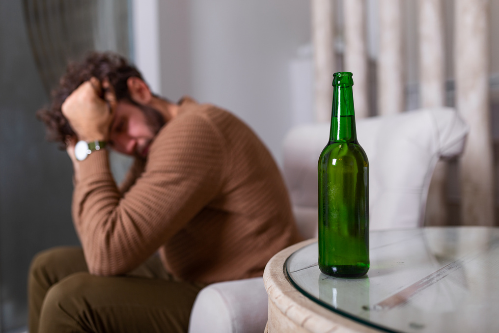 Silhouette of anonymous alcoholic person drinking behind bottle of alcohol. Man fighting with alcoholic habits, drink sitting on sofa at home